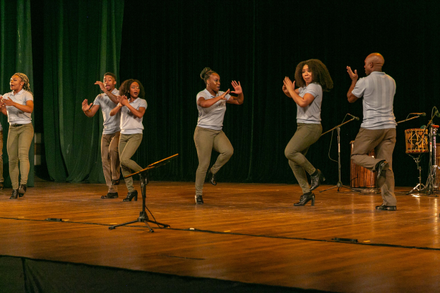 Eine Gruppe von Menschen auf einer Bühne mit Mikrofonen, Musikinstrumenten und einem Vorhang im Hintergrund, die Hände klappend in Feierlaune, die wie ein Tanzteam aussehen.