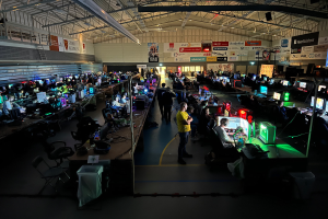 Ein voller Raum mit Menschen, die Videospiele spielen, sitzend und stehend, umgeben von Gaming-Equipment und Monitoren, mit Text und Logos auf Tafeln im Hintergrund.