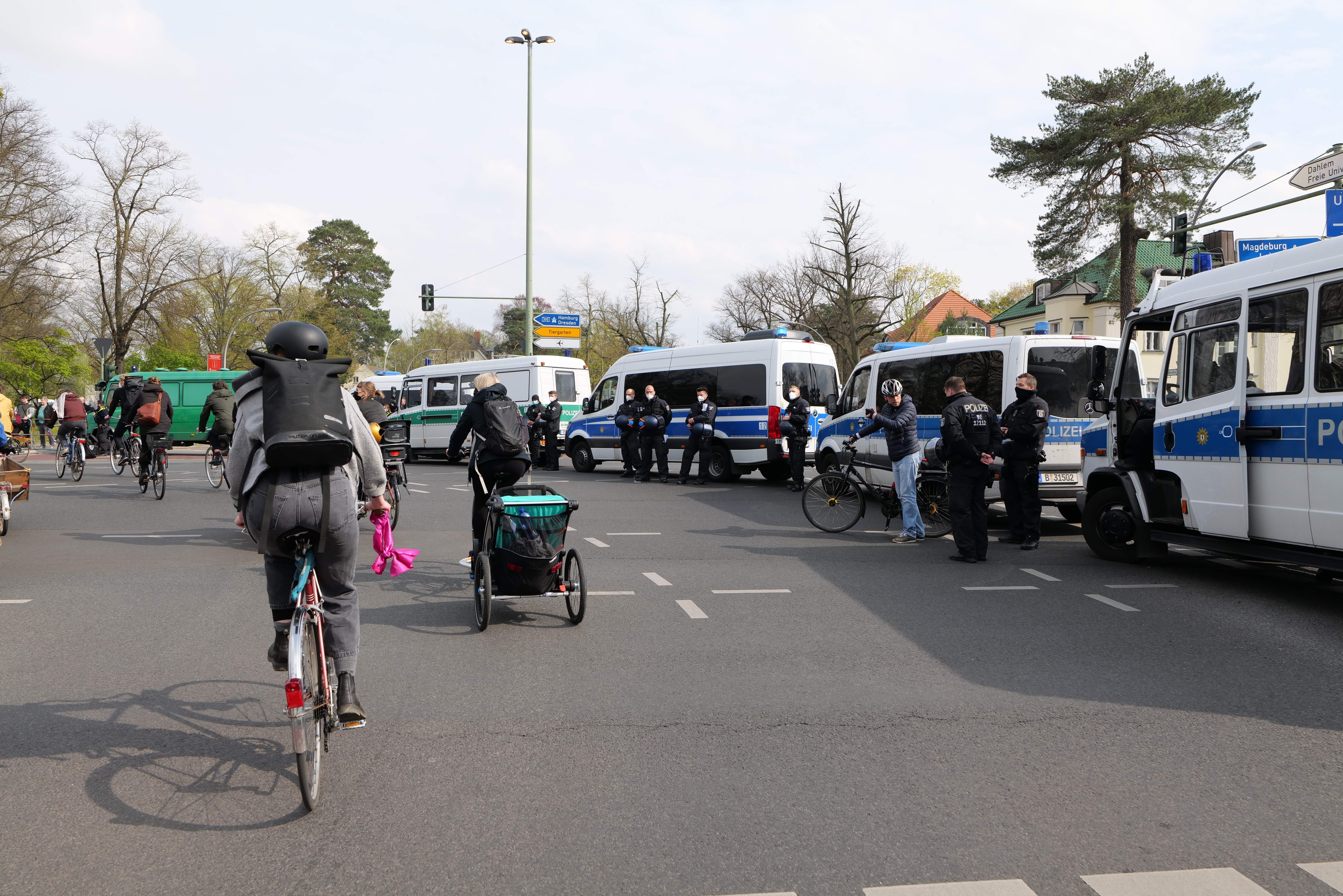 Eine Gruppe von Menschen, die auf Fahrrädern eine Straße entlangfahren, mit Polizeiwagen, anderen Fahrzeugen und städtischer Infrastruktur im Hintergrund.