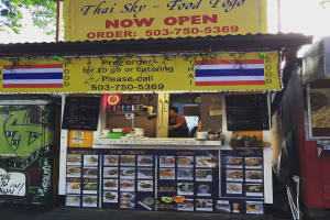 Ein Schaufenster mit verschiedenen Artikeln wie Getränkedosen, Speisekarten, einer Uhr, Gewürzen in Plastikbehältern, Kondimente, Werbetafeln und Namensschildern unter einem Himmel.