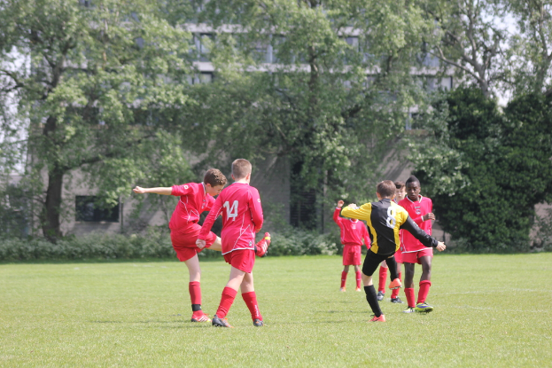 Fünf Jungs in roten T-Shirts und Shorts, die auf einem grünen Fußballfeld mit Bäumen und Gebäuden im Hintergrund Fußball spielen.