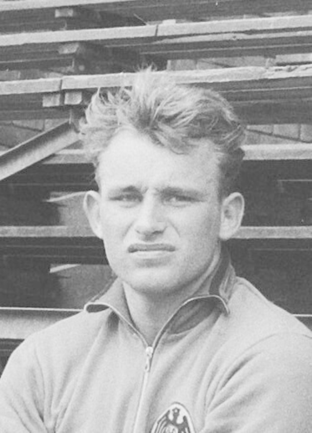 Schwarzes und weißes Foto eines Mannes in einer Jacke, der vor gestapelten Holzplanken steht und angeblich ein legendärer deutscher Fussballer ist.