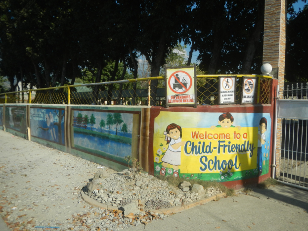 Ein Wandschild mit der Aufschrift "Willkommen in einer kinderfreundlichen Schule" umgeben von einem Metallzaun, einer Säule, Bäumen, Steinen und einem Weg.