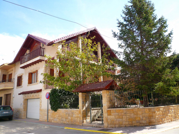 Ein Villa mit einem vorne geparkten Auto, das Fenster, Geländer, Säulen und ein Dach hat und von einem Tor, einem Schild, einem Straßenpfahl, Bäumen und einer Wand umgeben ist, unter einem bewölkten Himmel.