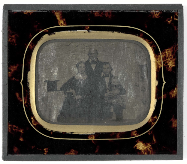 Schwarze-und-Wei├č-Foto eines Mannes in Anzug und Krawatte und einer Frau in einem unbekannten Outfit, die vor einem Fernseher stehen, beide mit ernsten Gesichtern.