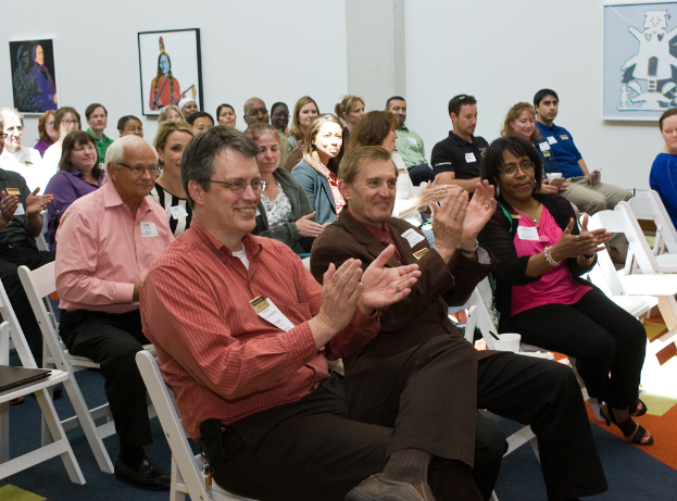Eine Gruppe von Menschen sitzt auf weißen Stühlen und klatscht, mit drei Foto Rahmen an einer weißen Wand dahinter und die Person in der Mitte trägt einen braunen Anzug mit Tags an ihrem Hemd.