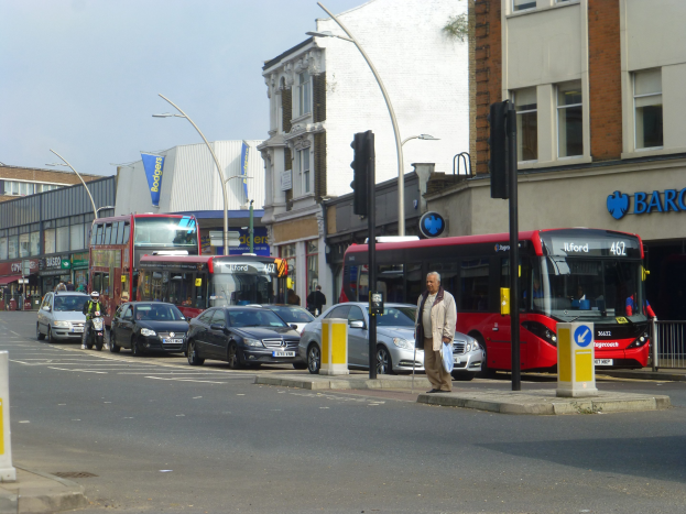 Ein roter Doppeldeckerbus fährt auf einer von Hochhäusern gesäumten Stadtstraße, umgeben von Passanten auf dem Gehweg, einem Radfahrer, Ampeln, Laternenmasten und Schildern an einem bewölkten Himmel.