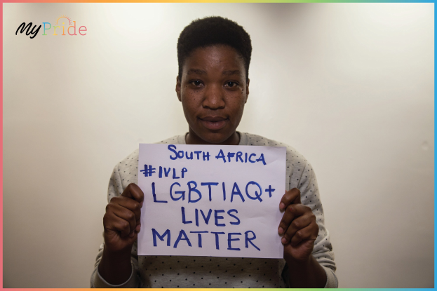 Eine Frau in einem weißen Hemd steht entschlossen vor einer Wand und hält ein Schild hoch, auf dem in fetten schwarzen Buchstaben 'LGBTQ+ Lives Matter' steht.