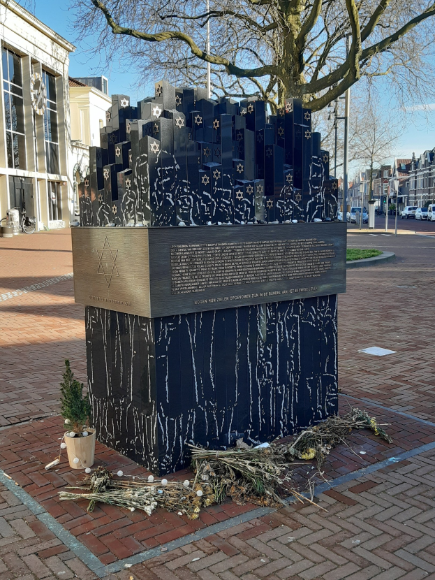 Denkmal in einem Stadtplatz für Holocaust-Opfer, mit einem Baum davor, einer Topfpflanze auf dem Boden, verstreuten trockenen Blättern und im Hintergrund Gebäude, Fahrzeuge, Masten und Bäume.