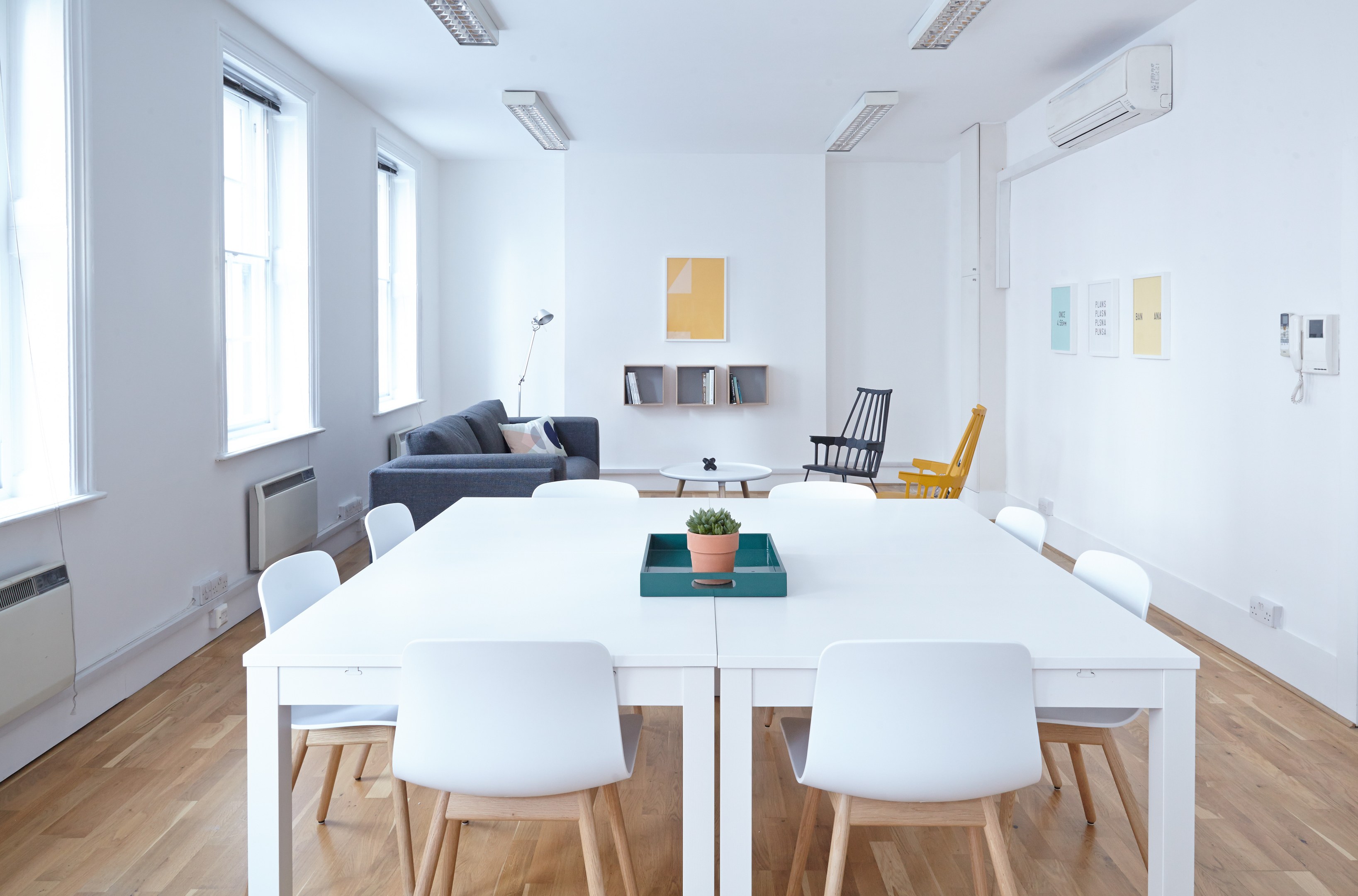 Innenansicht einer Wohnung in Berlin mit einem weißen Tisch mit einer Topfpflanze in einem Tablett, Stühlen, einem Sofa mit Kissen, einer Lampe, Regalen mit Gegenständen, einer Klimaanlage, Fotorahmen an der Wand, Fenstern, einer Decke mit Leuchten und einem Dach.
