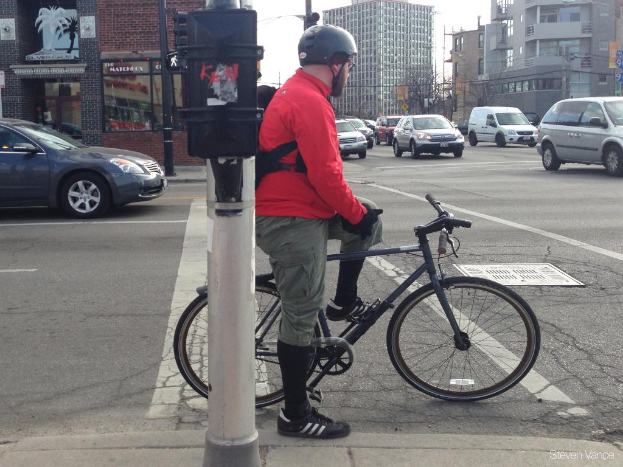 Eine Person sitzt auf einem Fahrrad in der Mitte des Bildes, mit ein paar Autos rechts, einem Auto links und Gebäuden im Hintergrund.