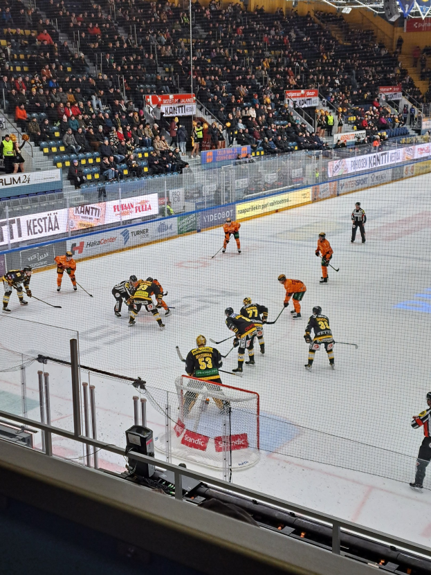 Eine Gruppe von Menschen, die Hockey auf einem Eisstadion spielt, mit einem Netz, einer Umzäunung, sitzenden Zuschauern, Bannern und Deckenbeleuchtung.