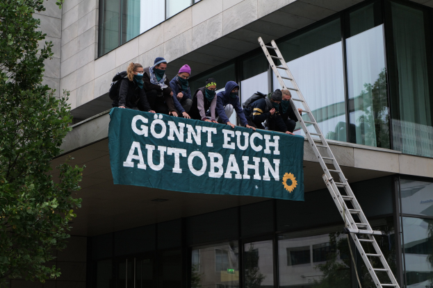 Gruppe von Menschen auf einem Gebäude, die eine Fahne halten, mit einem Baum links und Glasfenstern im Hintergrund, wahrscheinlich protestieren sie für die deutsche Regierung.