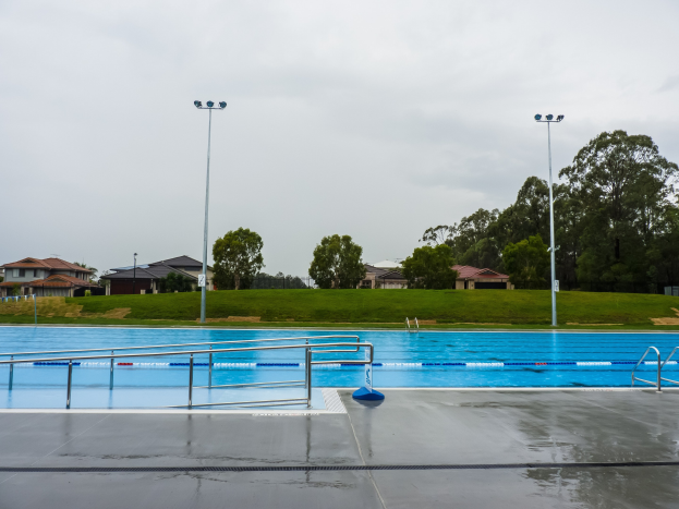 Großer rechteckiger Swimmingpool in einem Park, umgeben von Zaun, Pfählen und Laternen, mit Bäumen und Häusern im Hintergrund unter einem klaren blauen Himmel.