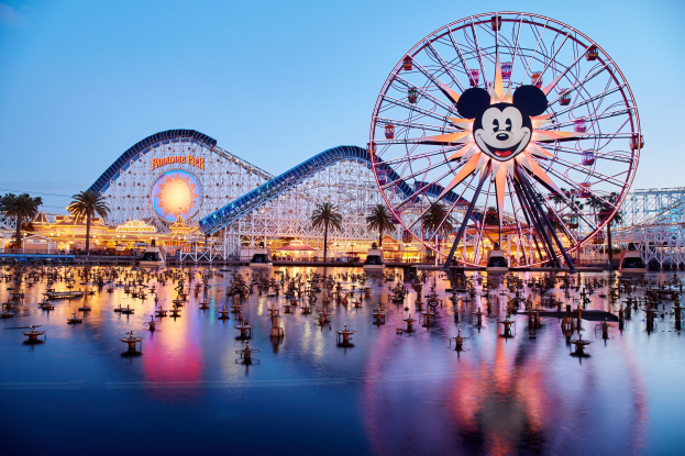 Eine lebendige Dämmerungsszene im Disneyland California Adventure mit einem riesigen Mickey-Maus-Rad im Vordergrund, einer Achterbahn im Hintergrund und Booten auf dem Wasser, mit einem Himmel aus Orangen, Pinks und Purpurtönen und silhouettenartigen Bäumen.