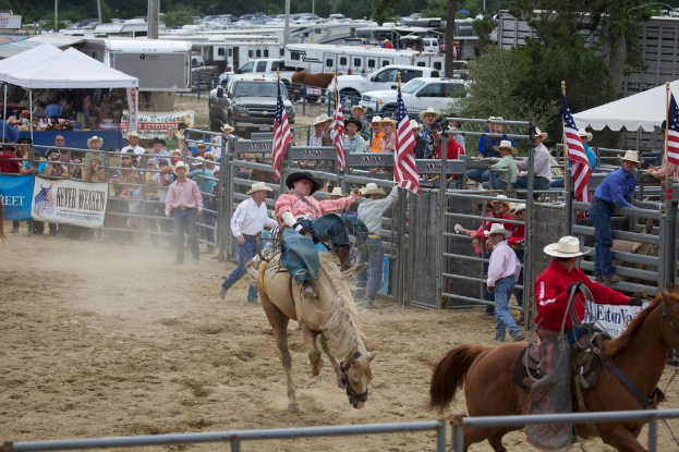 Zwei Personen reiten Pferde bei einem Wettbewerb namens Charreada, umgeben von Gruppen von Menschen, Bannern und Fahnen, Pavillonzelten, geparkten Fahrzeugen und Bäumen im Hintergrund.