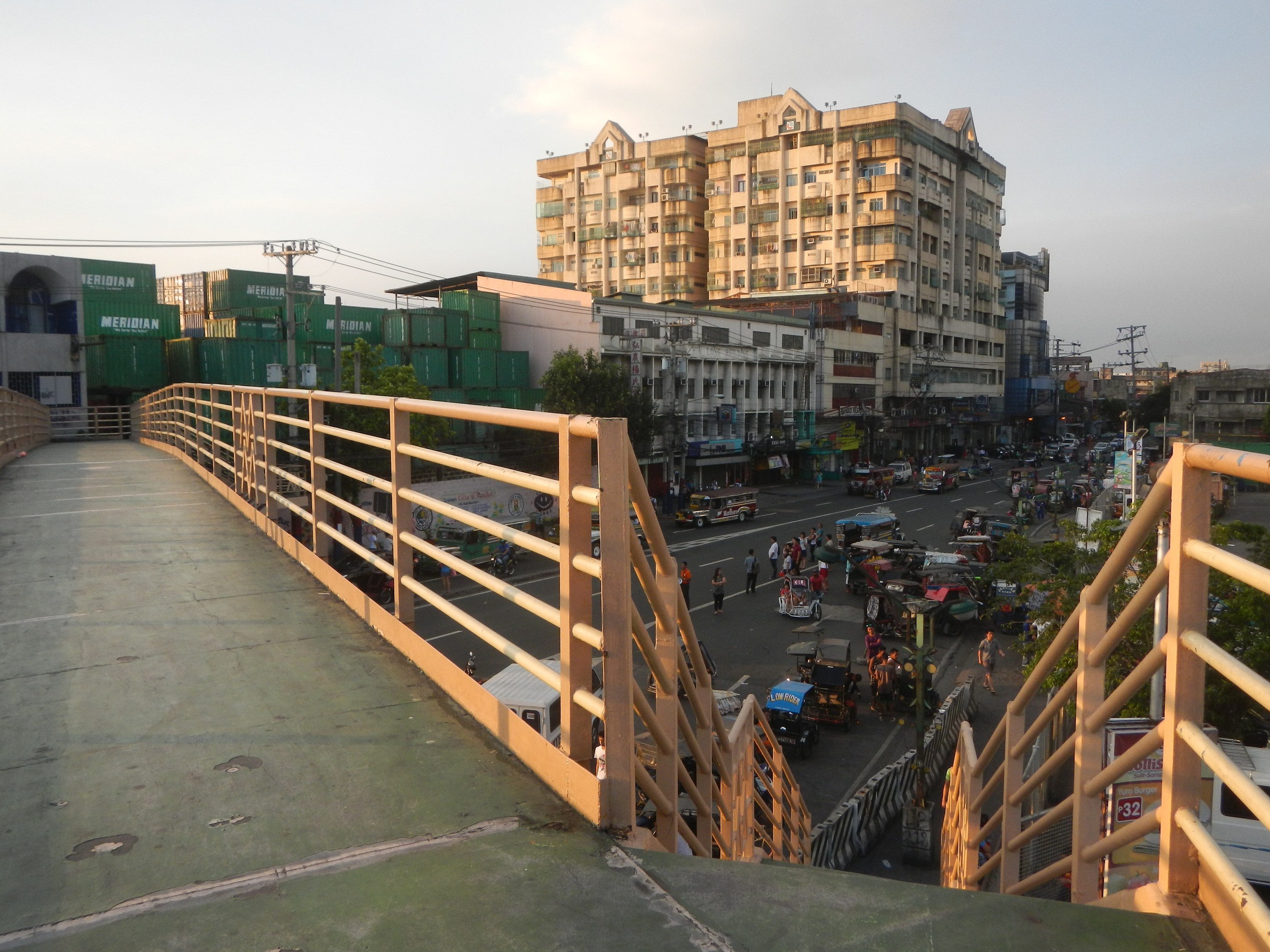 Eine Fußgängerbrücke mit Geländern überspannt eine vielbefahrene Straße mit Fahrzeugen und Fußgängern, im Hintergrund sind Gebäude, Bäume, Strommasten und ein bewölkter Himmel zu sehen.