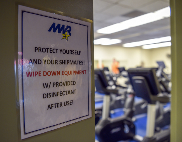 Ein Schild an einer Gym-Tür mit der Aufschrift "Schützen Sie sich und Ihre Schiffskameraden" mit Laufbändern und Deckenleuchten im Hintergrund, das Bild leicht unscharf, um Bewegung anzudeuten.