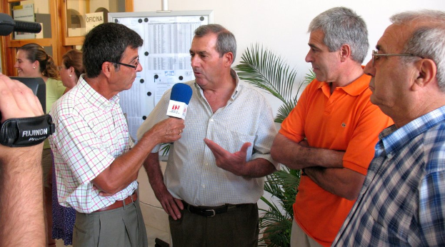 Ein Mann in der Mitte interviewt eine andere Person mit einem Mikrofon, während ein Mann links das Video mit einer Kamera aufzeichnet. Rechts stehen zwei Personen mit gefalteten Händen, und im Hintergrund gibt es eine Pflanze. Links neben der hölzernen Tür stehen zwei Mädchen, und in der Mitte zeigt ein Anschlagbrett Papiere.