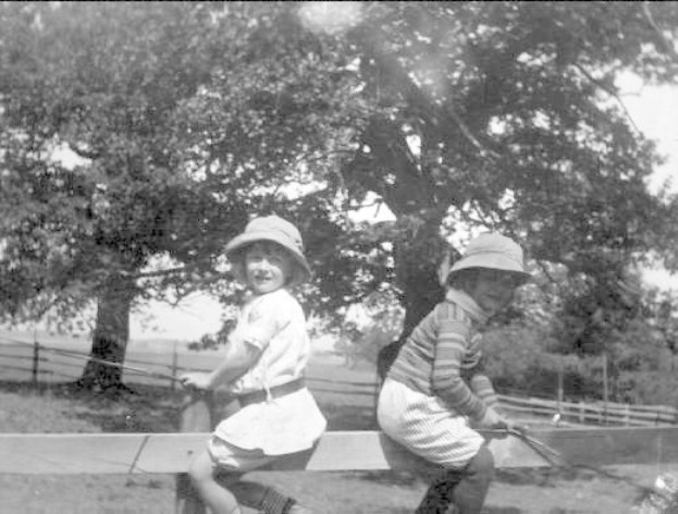 Zwei junge Jungs in Hüten halten Baseballschläger auf einem grasbewachsenen Feld mit Bäumen und einem Zaun im Hintergrund, in Schwarz-Weiß.