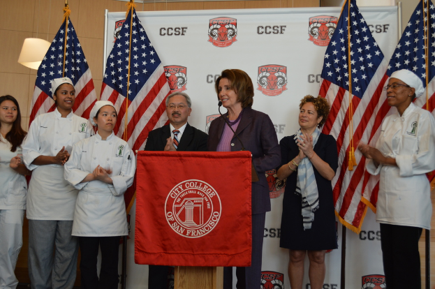 Eine Frau steht an einem Podium mit einem Mikrofon, lächelt, vor einem roten Tuch, Fahnen, einer Fahne, einer Lampe und einer Wand im Hintergrund.
