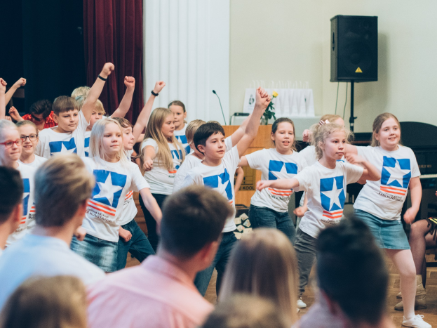 Kinder in weißen Hemden und blauen Shorts tanzen vor einem sitzenden Publikum bei einem internationalen Schul-Tanzwettbewerb, mit einer Vorhangwand, Lautsprecher und einem Tisch mit Mikrofon im Hintergrund.