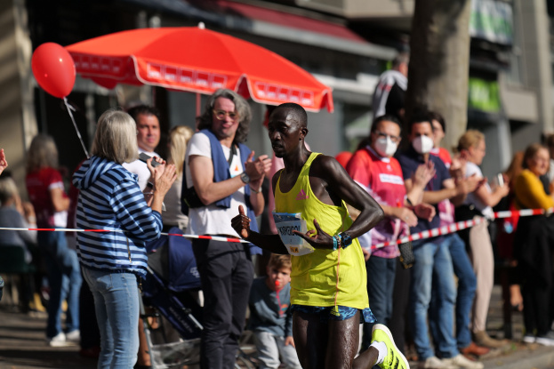 Ein Mann in einem gelben T-Shirt läuft bei einem Marathon, umgeben von einer jubelnden Menge mit einem Schirm und einem Kinderwagen im Hintergrund.