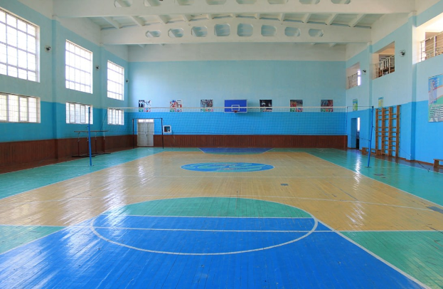 Große Turnhalle mit blau-weißem Boden, Basketballfeld, Netz mit Pfosten, Tür, Fenstern, Wandplakaten, Geländer und Dach.