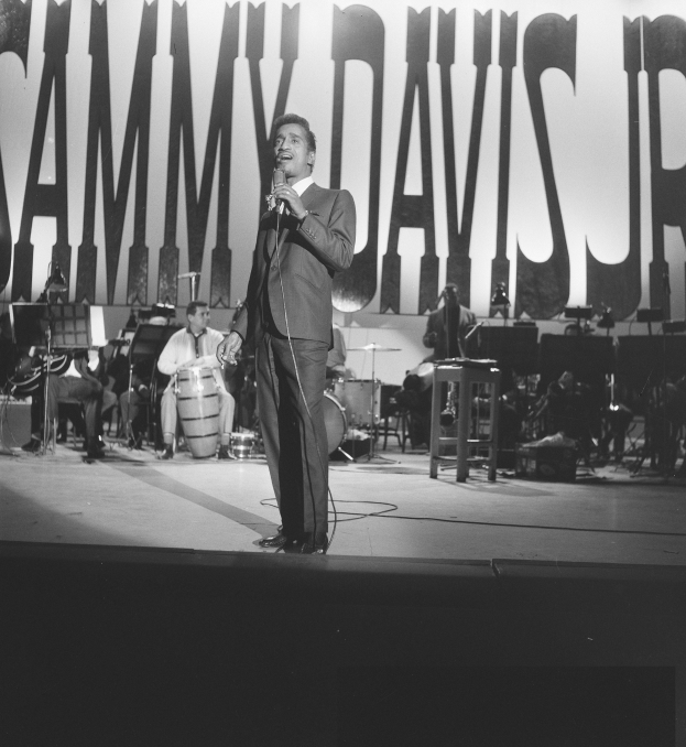 Schwarz-weiß-Foto von Sammy Davis Jr. auf der Bühne mit einem Mikrofon in der Hand, umgeben von Musikern.