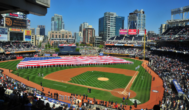 Baseballstadion voller Zuschauer bei einem Spiel, mit einer Gruppe unter einer amerikanischen Flagge auf dem Feld, Anzeigetafeln auf beiden Seiten und Gebäuden, Bäumen und einem klaren Himmel im Hintergrund.