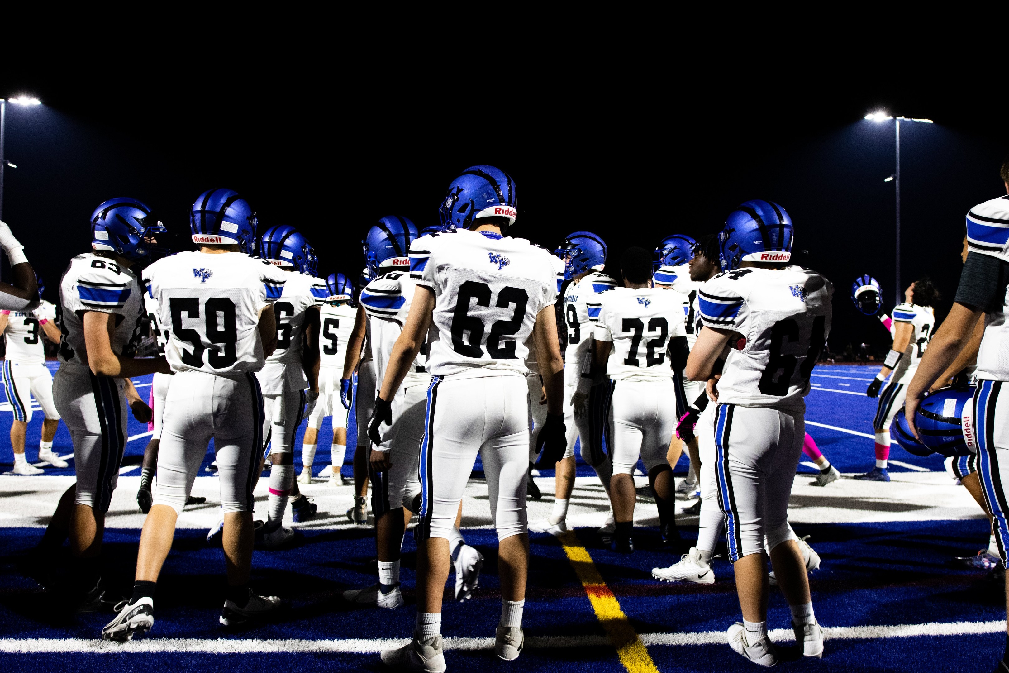 Eine Gruppe von Fußballspielern in Helmen und Stiefeln steht auf einem Feld unter Flutlicht, bereit für ein Spiel.
