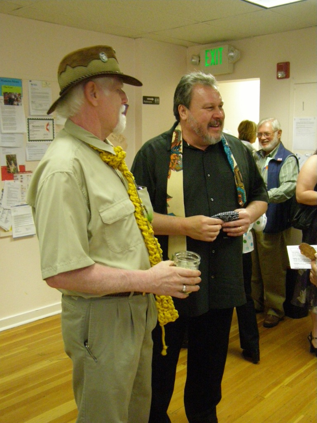 Eine Gruppe älterer Männer steht auf einem Holzboden in einem Raum mit Papieren an den Wänden, einer Warntafel an der rechten Wand, einer Tür darunter und Deckenlampen.
