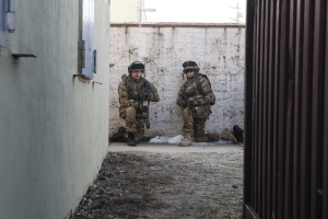 Zwei Soldaten stehen in der Mitte des Bildes, umgeben von Erde, mit einer Wand und einem Fenster auf der linken Seite, einer Eisenwand auf der rechten Seite und Gebäuden, einem Strommast und einer weiteren Wand im Hintergrund.