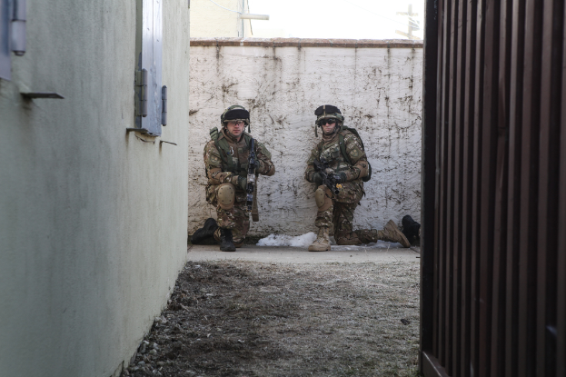 Zwei Soldaten stehen in der Mitte des Bildes, umgeben von Erde, mit einer Wand und einem Fenster auf der linken Seite, einer Eisenwand auf der rechten Seite und Gebäuden, einem Strommast und einer weiteren Wand im Hintergrund.