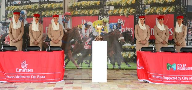 Sechs Flugbegleiterinnen in braunen Anzügen stehen lachend und posend vor der Kamera, mit einem Emirates-Tisch im Vordergrund und einem Polo-Plakat im Hintergrund, während eine goldene Trophäe auf einem weißen Tisch in der Mitte steht.