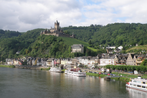 Ein malerischer Blick auf den Rhein in Deutschland, mit einer Burg auf einem Hügel, Booten auf dem Fluss und Fahrzeugen auf der Straße.