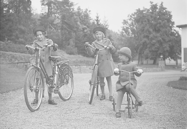 Drei Kinder in Mänteln und Mützen fahren Fahrräder in einem Park mit Bäumen und einem Haus im Hintergrund, in Schwarz-Weiß.