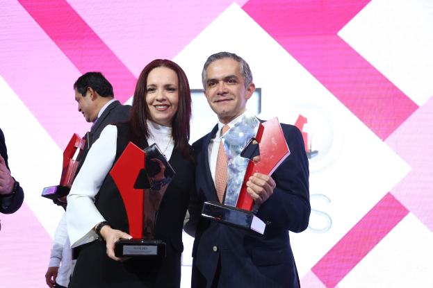 Zwei strahlende Individuals mit Trophäen auf einer Bühne bei einer Preisverleihung, mit einem sichtbaren Bildschirm im Hintergrund.