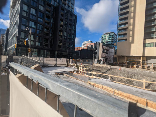 Baustelle in Toronto mit einem großen Gebäude im Bau, umgeben von Absperrungen, Pfählen, Verkehrssignalen und Schildern unter einem bewölkten Himmel.
