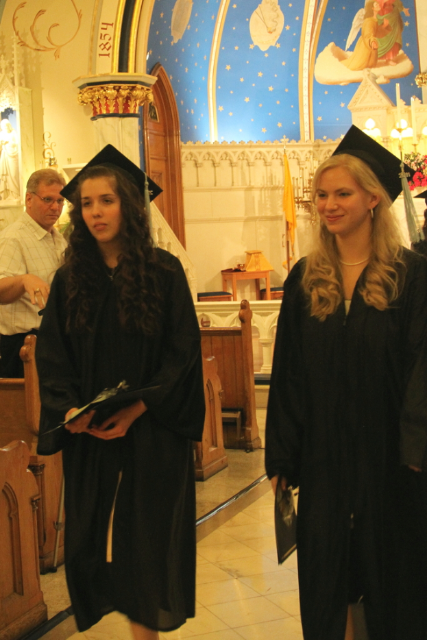 Zwei Frauen halten Papiere oder Zertifikate, eine davon lächelt; ein Mann steht links neben Bänken, und im Hintergrund ein Tisch mit Hockern, eine gelbe Flagge, Treppe, Säule, Tür und weiße und blaue Wände.