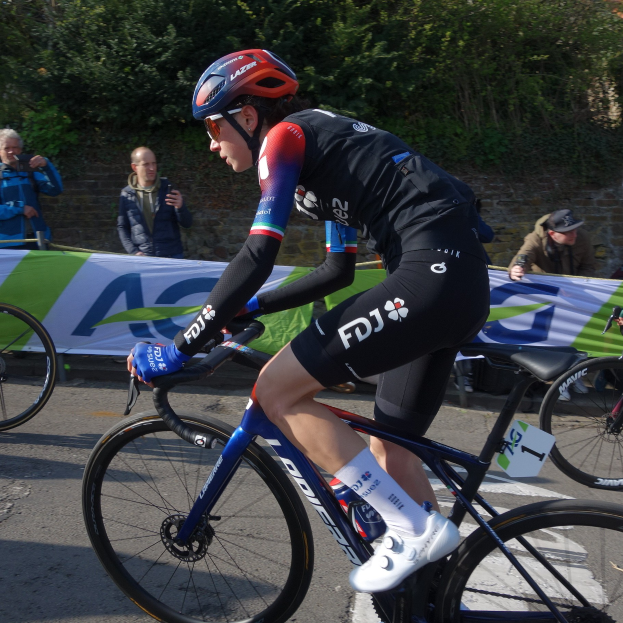 Die professionelle Radrennfahrerin Katie Archibald trägt einen Helm, eine Schutzbrille, Handschuhe und Schuhe und fährt ein Fahrrad auf einer Straße an einer Zuschauermenge vorbei, mit einem Banner, einer Wand, Bäumen und einem Gebäude im Hintergrund.