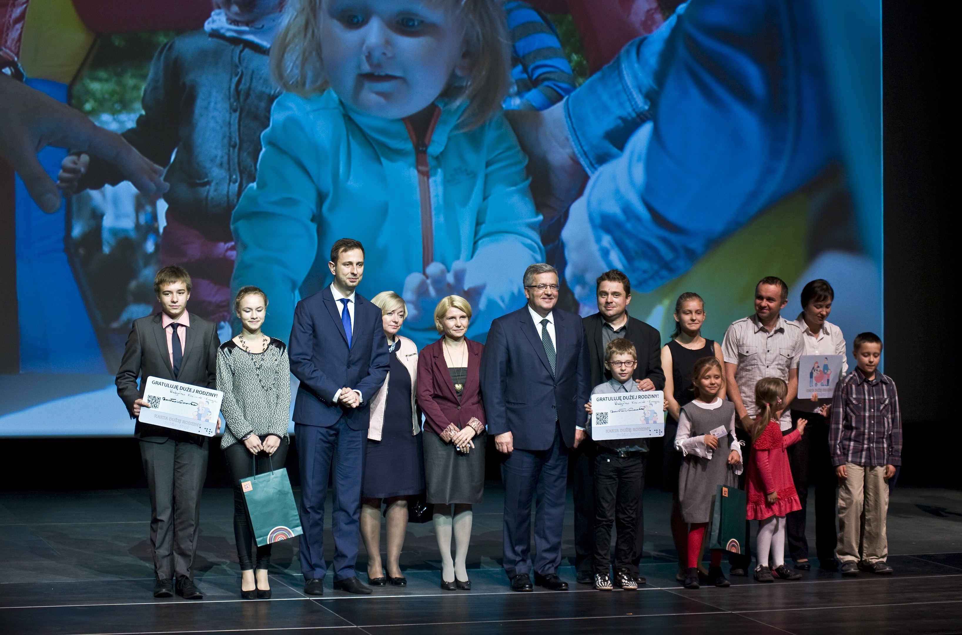 Eine Gruppe von Menschen auf einer Bühne mit Taschen und Urkunden, im Hintergrund ein Bildschirm mit einem Bild von einem Kind und anderen.