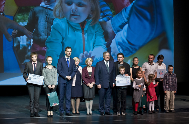Eine Gruppe von Menschen auf einer Bühne mit Taschen und Urkunden, im Hintergrund ein Bildschirm mit einem Bild von einem Kind und anderen.