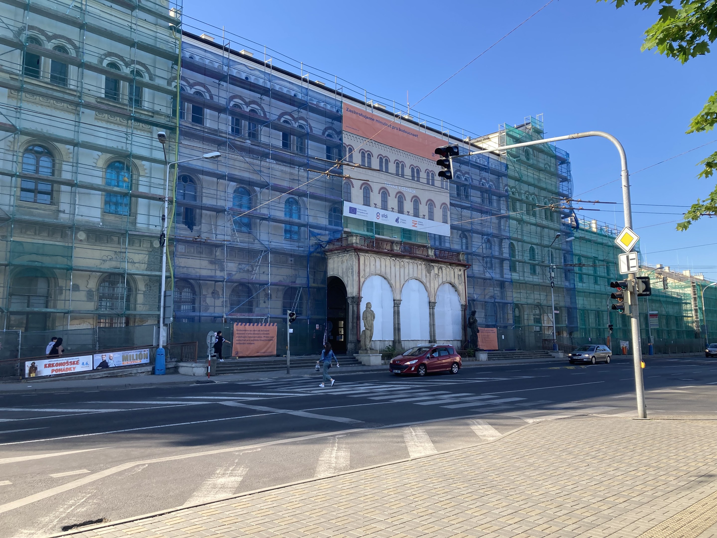 Ein großes Gebäude mit Gerüst, das renoviert wird, umgeben von Fahrzeugen, Passanten, Ampeln, Hinweistafeln, Straßeninfrastruktur, Bäumen und einem klaren blauen Himmel.