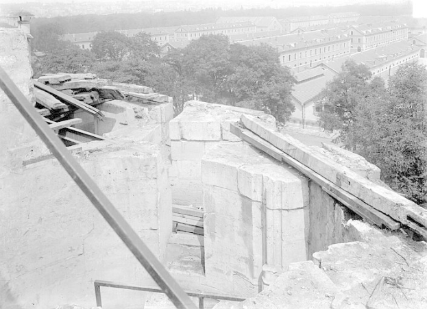 Ein Schwarz-Weiß-Foto eines teilweise abgerissenen Gebäudes mit verstreuter Trümmern, Bäumen und anderen Gebäuden im Hintergrund.