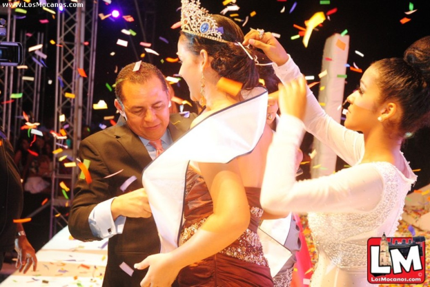 Ein Mädchen mit einer Krone auf der Bühne eines Miss-World-Events, hinter ihr eine andere Frau, die die Krone anpasst, und ein Mann im schwarzen Anzug, der vor ihr die Hand schüttelt, vor einem Hintergrund aus Metallrahmen und dunkler Kulisse.