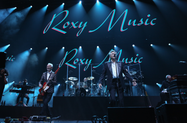 Eine Gruppe von Musikern, die auf der Bühne mit Instrumenten, Mikrofonen und Ständern spielen, mit Kabeln und Equipment vorne und einem Schild im Hintergrund, auf dem "Roxy Music - Live in der O2 Arena, London" steht.