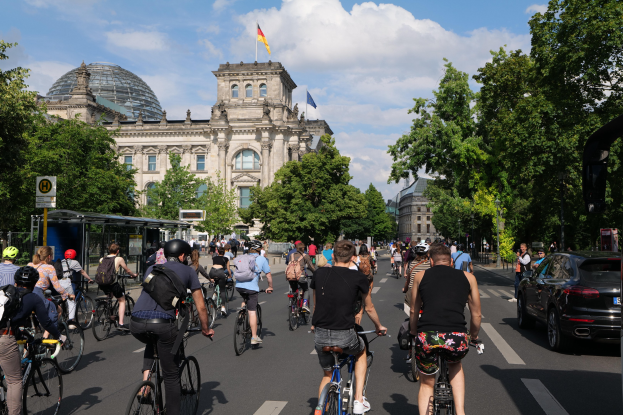 Eine Gruppe von Menschen, die auf Fahrrädern eine von Bäumen gesäumte Straße in Berlin, Deutschland, entlangfahren, mit Gebäuden und einer Bushaltestelle im Hintergrund, unter einem bewölkten Himmel und einer Flagge auf einem der Gebäude.