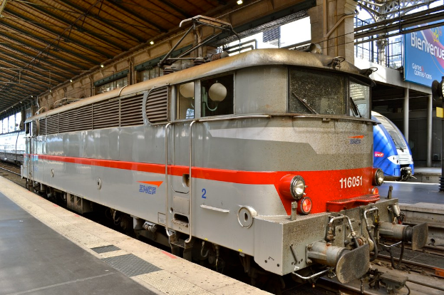 Eine Lok auf Schienen im Vordergrund mit einer Plattform links und einem Schuppen darüber, mit einem weiteren Zug, einer Plattform, einem Banner und Pfählen im Hintergrund.