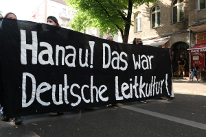 Eine Gruppe von Menschen, die eine Straße entlanggehen und ein schwarzes Banner mit der Aufschrift 'Hanau das war deutsche Leitkultur' tragen, mit Gebäuden, Bäumen und einem klaren blauen Himmel im Hintergrund.